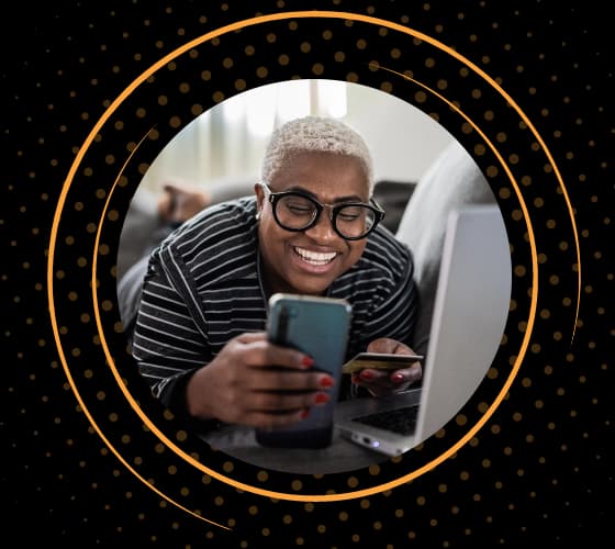 Persona sonriendo mientras mira un smartphone con un portátil abierto delante.