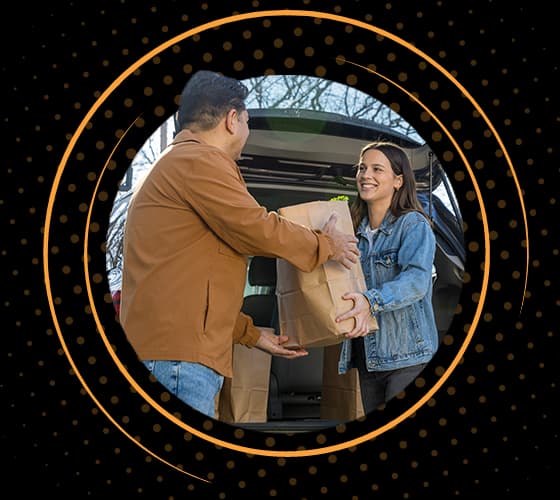 Una mujer sonriente entrega una bolsa de la compra a un hombre con chaqueta marrón desde la parte trasera de un todoterreno.